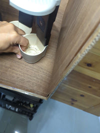 A close-up view of a hand holding a small beige plastic cup or container under a dark metal and white device on a wooden surface.